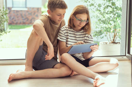 Teenage siblings children reading using tablet at home in summer sunny dayの写真素材