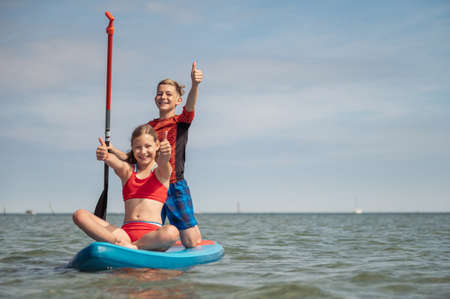Happy teenage siblings playing and having fun on paddle board at sunny summer day at vacation on seaの写真素材