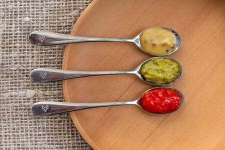 Adjika sauce, mustard and pesto in spoons on wooden plate on burlap. Top view, photo of food, wooden...
