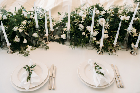 Decorated tables with plates, knives, forks and bouquet with white flowers and greens on the centreの写真素材