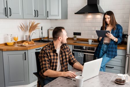 Beautiful couple of freelancers working from home. A guy sits at a table with a laptop, a girl stands with a tablet in a kitchenの写真素材