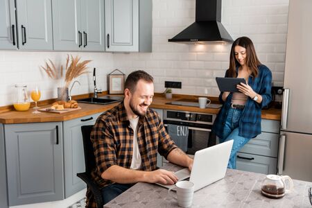 Beautiful couple of freelancers working from home. A guy sits at a table with a laptop, a girl stands with a tablet in a kitchenの写真素材