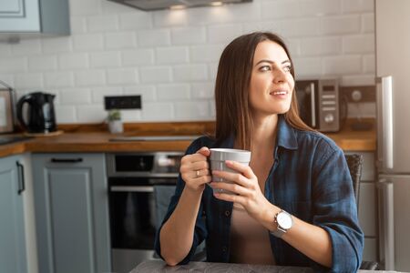 A young girl sits at a table with a gray cup in her hands and looks out the windowの写真素材