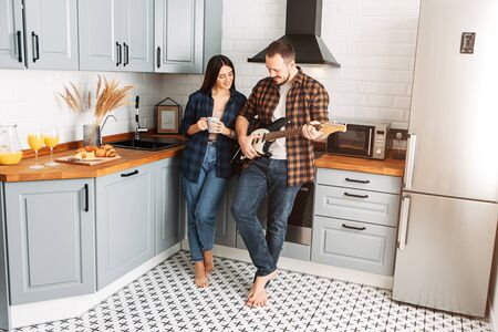 A guy with a beard and mustache in the kitchen with an electric guitar. Near a beautiful brunette girl with a cup in her handsの写真素材