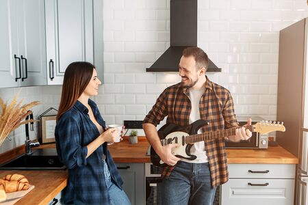 Couple in casual clothes at home in the kitchen. Guy with electric guitar, girl with coffeeの写真素材