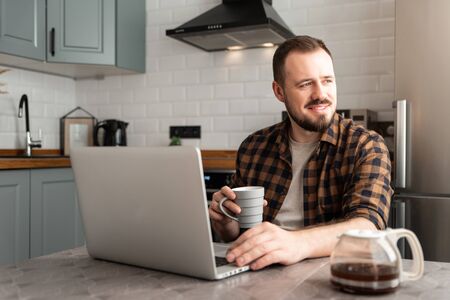 A guy in a casual shirt with a cup in his hand sits with a laptop and laughs