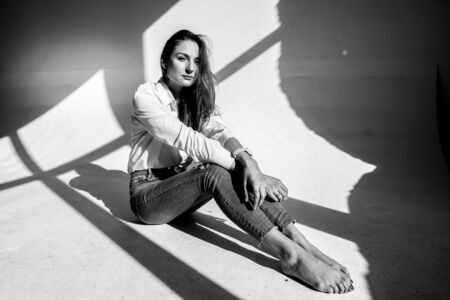 Slender beautiful girl in jeans and a white shirt sits on the floor in the studio. Shadows from the window frame in the backgroundの写真素材