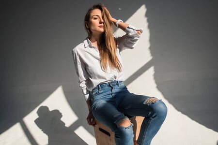 A girl in a white shirt and jeans holds a hand in her hair. She is sitting on a wooden cube in the studioの写真素材