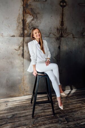 Girl in a business white suit in the studio in the loft style. She is sitting in a high chair and laughingの写真素材