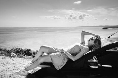 Beautiful girl lying on a car under sunlights, sea on backgroundの写真素材