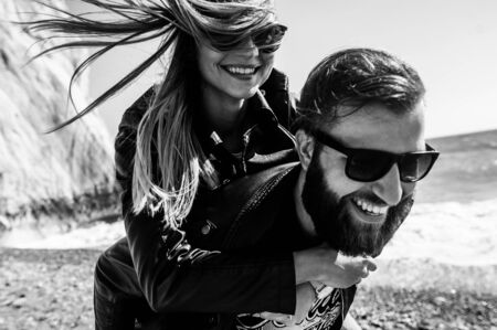 Black and white photo of happy couple in the leather jackets running on the beach. They are in sunglasses. A guy with a beard.の写真素材