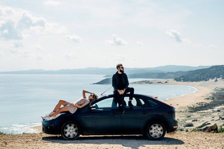 Couple sitting on a car, sea and mountain on background