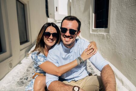 Happy couple hugging and laughing together, sitting on a narrow street with a view of Santoriniの写真素材