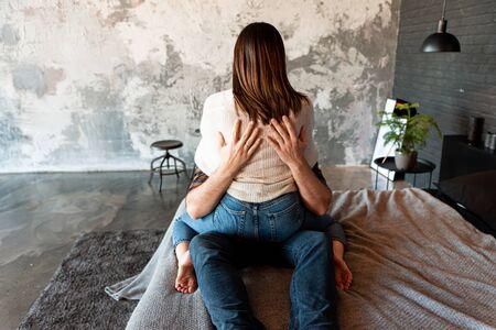 Beautiful young couple hugs sitting face to face on a large bed, back viewの写真素材