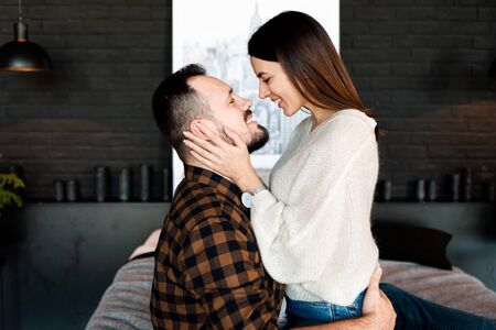 Beautiful young couple hugs sitting face to face on a large bed, side viewの写真素材