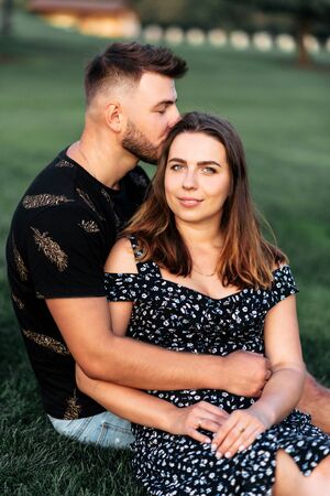 Summer love story. Portrait of a couple in love outdoors on the grass. A handsome guy with a beard gently hugs and kisses his beautiful girlfriendの写真素材