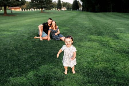 Family summer walks in the park. Little baby girl runs barefoot on the grass, her young parents are sitting in the backgroundの写真素材