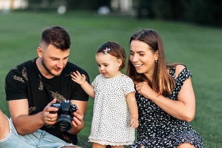 Family outdoors in the summer. Young parents spend time with their baby girl in a park. Dad shows his daughter the pictures on the screen of his cameraの写真素材