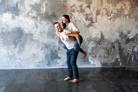 Young attractive couple in love having fun in a loft room. A guy with a beard carries a girl on his back. They are in white t-shirts and jeans.の写真素材