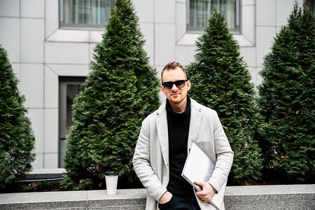 Positive attractive guy freelancer with laptop outdoors. He is wearing a stylish smart casual and sunglasses.の写真素材
