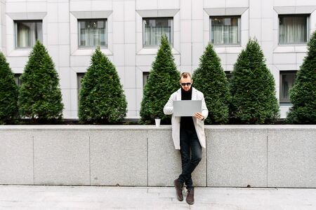 Positive attractive guy freelancer with laptop outdoors. He is wearing a stylish smart casual and...