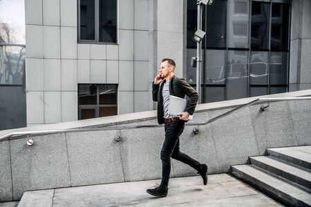 Time is running out. A young man in chic casual clothes runs up the stairs to the business center of the city. He is talking on the phone, in his hands is a laptop.の写真素材