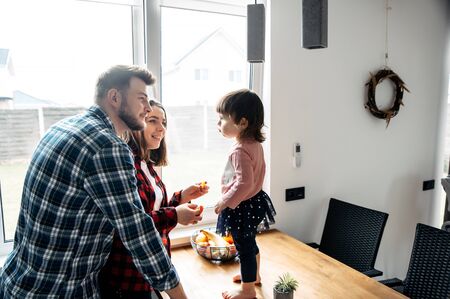 Young family in the kitchen. A little girl stands in front of her parents and eats fruit, parents...