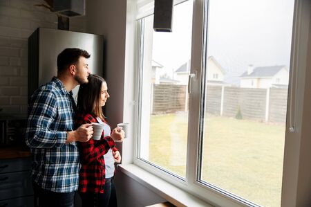 Love is in the air. Couple in love hugging in the kitchen at home. They look out the window with coffee mugs in their hands.の写真素材