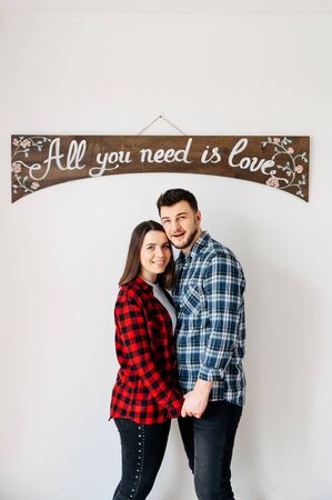 Love story. Young and in love guy and a girl in casual shirts in a checked box holding hands under a sign all you need is love. Indoor picture on a white backgroundの写真素材