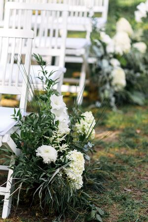 Outdoor wedding ceremony. White wooden chairs in a row on the grass, decorated with white fresh flowers and green branches.の写真素材