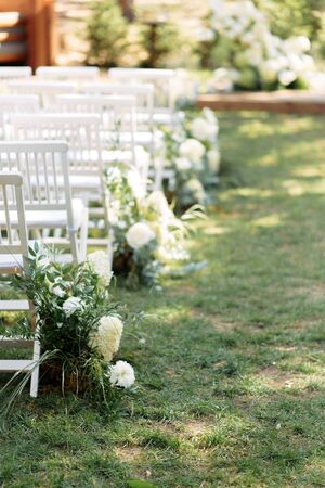 Outdoor wedding ceremony. White wooden chairs in a row on the grass, decorated with white fresh flowers and green branches.の写真素材