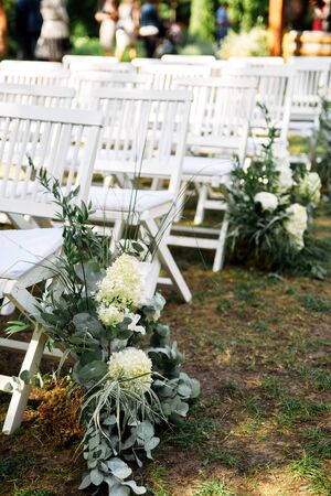Outdoor wedding ceremony. White wooden chairs in a row on the grass, decorated with white fresh flowers and green branches.の写真素材