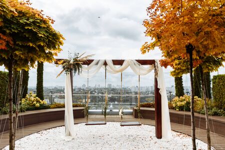 Autumn wedding ceremony outdoors. Roof swing with city view decorated with flowers and white fabricの写真素材