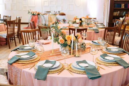 Round festive table in peach and green colors indoors. Elegant table setting with fabric napkins on plates, bouquets of fresh flowers in the center of the tableの写真素材