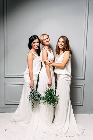 Bridesmaids are dressed in the same style. Three attractive young girls in long elegant dresses in a studio with gray wallsの写真素材