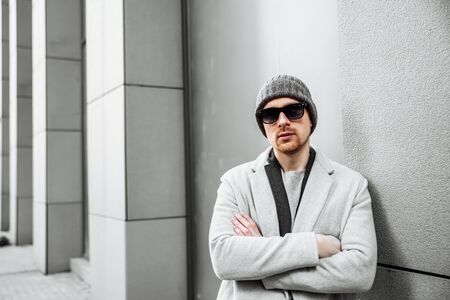 A young and attractive man with a beard and in sunglasses stands with his arms crossed over his...