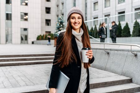 First day at work. A young woman with a laptop and coffee in her hands is standing near the office building and smiling.の写真素材