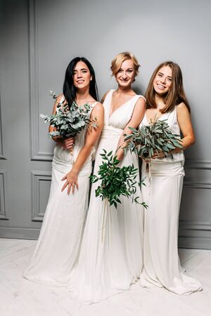 Boho style wedding. Bridesmaids in long ivory dresses, in their hands green bouquets. Photo in studio on a gray backgroundの写真素材