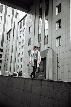 Stylish man in autumn winter clothes is walking around the city. He is wearing a coat, hat and sunglasses. High-rise concrete buildings in the backgroundの写真素材