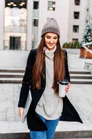 Young attractive girl student, freelancer in casual clothes urban landscape. Autumn season. She holds takeaway coffee and a laptop in her hands. Free Work Schedule Conceptの写真素材
