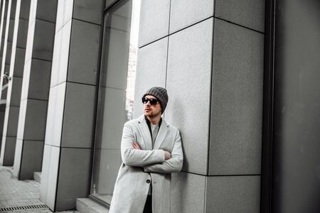 A young and attractive man with a beard and in sunglasses stands with his arms crossed over his chest. He is wearing a coat and hat. Horizontal pictureの写真素材