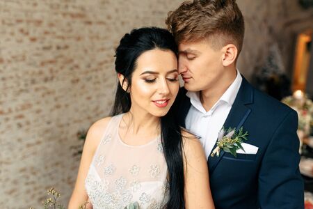 The happiest day. Portet of a newlywed couple in the studio in natural light. Groom in elegant suit gently hugs a beautiful bride with a closed eyesの写真素材