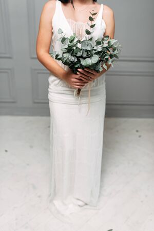 Bride or bridesmaid in long dress in studio on gray background with a nontrivial bouquet of green branches in her hands. Face is not visibleの写真素材