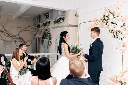 Indoor boho style wedding arch. A couple of newlyweds in front of the altar hold hands and look at each other with love. Guests are sitting on chairsの写真素材