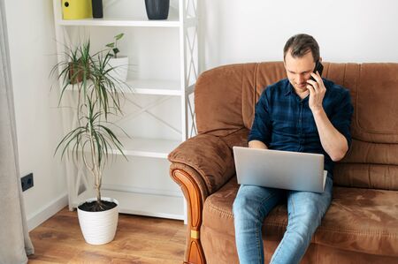 Home leisure. The guy is sitting at home on the sofa, talking on the phone and using a laptop. Online shopping, web surfing, social networksの写真素材