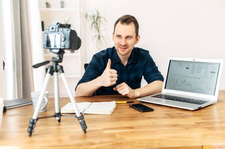 Put your thumb up. Young attractive man records blogs, video tutorials, training on camera. Camera on tripod in front of guyの写真素材