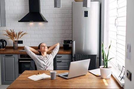 Home office, distant work, freelancer. Young woman takes a break, resting leaning back in a chair in front of a laptop in the kitchen at homeの写真素材