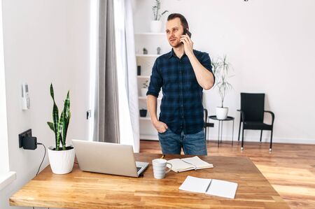 Office work. Young casual guy in a minimalistic modern office working using laptop. He is talking on a cell phone.の写真素材