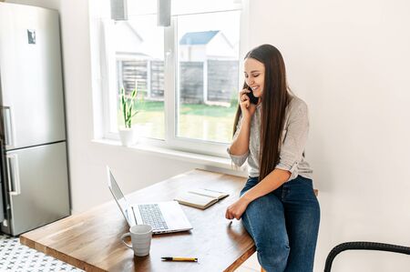 Work remotely, freelance, work at home. Young positive attractive woman relaxedly sits on the edge of the table and speaks on the phone, she looks at the laptop screen and smilesの写真素材