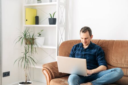 Home leisure. The guy is sitting at home on the sofa and using a laptop. Online shopping, web surfing, social networksの写真素材
